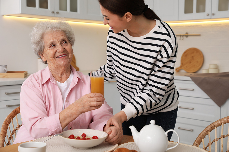 Young Caregiver Serving Breakfast For Senior Woman At Table In K Family visiting older parent receiving in-home senior care.