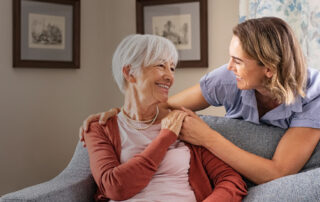 An older adult with short gray hair sits in a cushioned chair in a living room while another adult stands beside the chair with one arm resting around the seated person’s shoulders.
