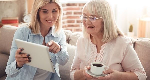 Smiling younger woman shows smiling older woman something on a tablet.