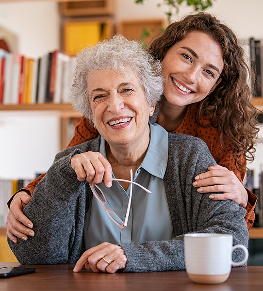 Young woman embracing senior mother at home and looking at camera. Portrait of happy adult granddaug