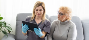 A blonde woman in grey scrubs shows another older blonde woman with glasses something on her tablet screen.
