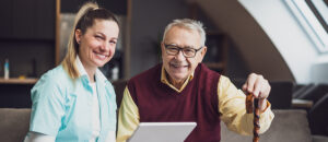 A blonde caregiver and an older gentleman in a maroon sweater smile over an electronic tablet.