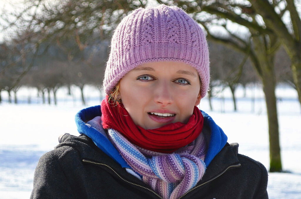 A woman wears a pink hat, a black coat with a blue lining, and red and white scarves in front of a snowy landscape.