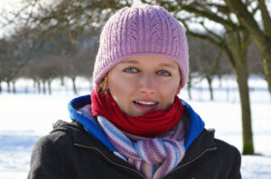 A woman wears a pink hat, a black coat with a blue lining, and red and white scarves in front of a snowy landscape.