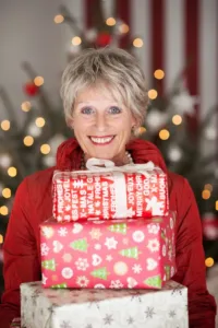 An older woman with blonde hair in a red coat holds a stack of wrapped presents.