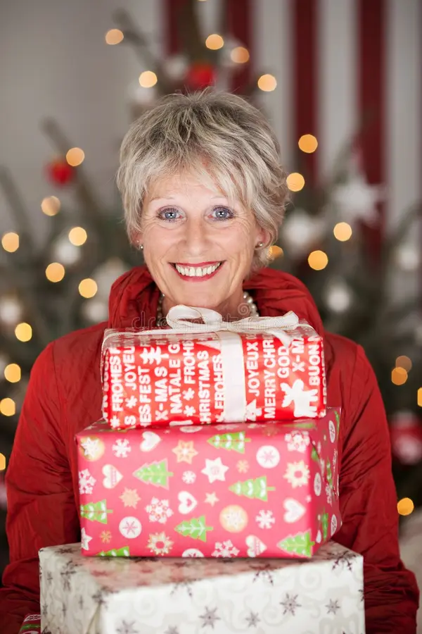 An older woman with blonde hair in a red coat holds a stack of wrapped presents.