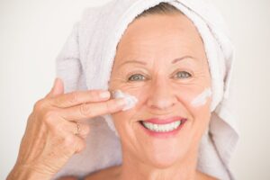 An older women with her hair in a white towel slathers cream on her face with a smile.