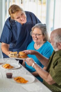 An older woman in a cerulean shirt sits between a standing woman in a dark blue shirt and a man in an olive green shirt. The standing woman in handing the man a sandwich. 