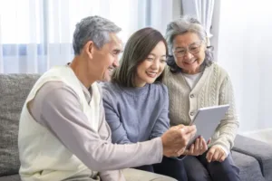 Two older people sit on either side of a young woman in a blue sweater and they are all looking at a tablet and smiling.