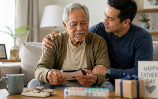Two people sit together on a living room couch, looking at a small photo or card. The older individual holds the item while the younger individual rests an arm across the older individual’s shoulders. A table in front displays a mug, eyeglasses, a notebook, a weekly pill organizer, and a wrapped gift. A greeting card on the table reads “Happy Father’s Day.”