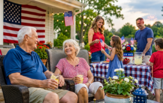Several people gather in a backyard decorated with American flags. Two older individuals sit in outdoor chairs in the foreground holding drinks, while others stand nearby, including a child holding a small flag. A table covered with a red, white, and blue checkered cloth holds drinks and decorations, and a house with a large American flag is visible in the background.