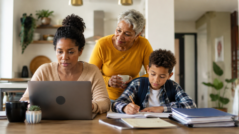 Three generations of a family gathered around a table at home, with one person working on a laptop, another looking on, and a child completing school assignments.