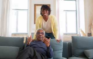 An older adult sits on a sofa holding a smartphone and giving a thumbs-up gesture, while another adult stands behind the sofa in a bright, modern living room with large windows.