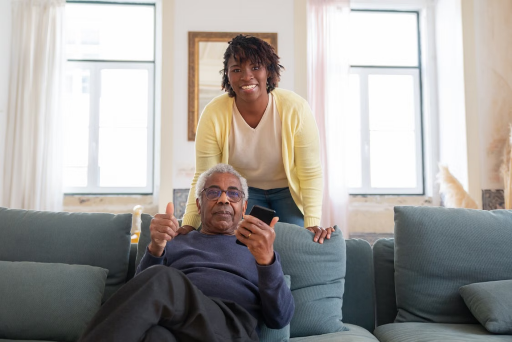 An older adult sits on a sofa holding a smartphone and giving a thumbs-up gesture, while another adult stands behind the sofa in a bright, modern living room with large windows.
