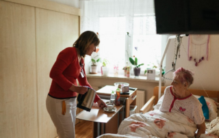 Caregiver serving a drink and providing assistance to an older adult sitting up in bed at home, illustrating in-home senior care support.