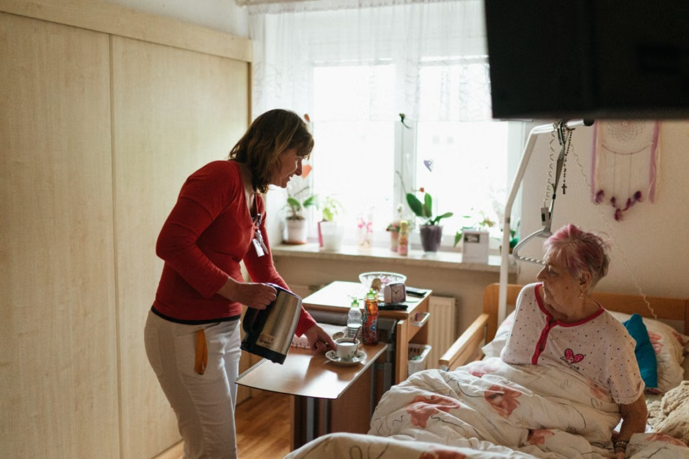 Caregiver serving a drink and providing assistance to an older adult sitting up in bed at home, illustrating in-home senior care support.